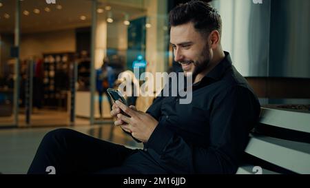 Caucasian stylish man in black suit successful businessman customer sit at shopping mall using mobile phone apps make online order buying clothes win Stock Photo