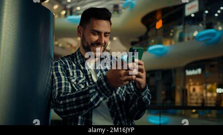 Young man blogger arabian hispanic businessman stand in shopping center look at mobile screen texting message email read positive news on cellphone Stock Photo