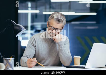 Serious thinking architect designer working on blueprint, mature man working inside office engineer designing on whatman. Stock Photo