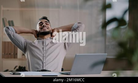 Happy satisfied arab man worker businessman hispanic guy finished work task on computer sit at home office put hands behind head lean on chair take re Stock Photo