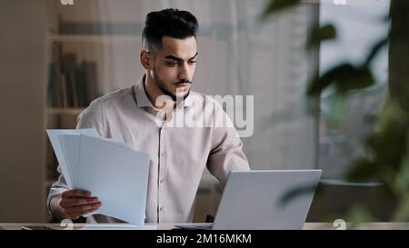 Serious arabian young business guy male professional worker employee type on computer keyboard copy paper text at electronic form focused hispanic man Stock Photo