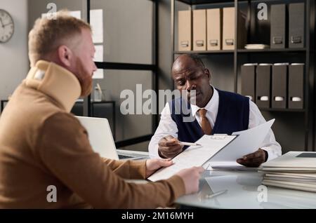 Portrait of black insurance broker helping man with neck brace filling legal forms after accident Stock Photo