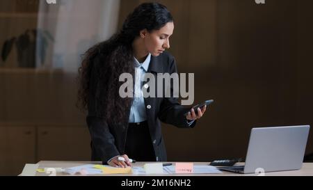 Focused female arabian employee designer business woman standing at office desk using mobile phone checking information writing tasks on sticky notes Stock Photo