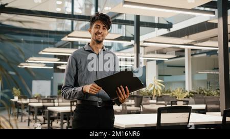 Young arabian confident business man guy stand in modern office hold folder check document male hispanic entrepreneur accountant read financial report Stock Photo