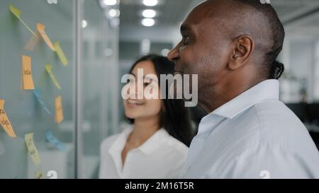 Corporate team business partners colleagues look at paper memo stickers on wall discuss project giving business advice managing tasks structure sticky Stock Photo