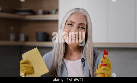 Playful woman housewife elderly caucasian granny janitor wears yellow rubber gloves posing with wet sponge and sprayer splashing detergent in air havi Stock Photo