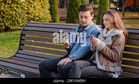 Caucasian couple two young people partners sit on bench use phone cell gadgets scroll internet web pages check social networks show images each other Stock Photo