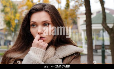 Doubtful pensive young woman brunette caucasian girl stand alone in autumn urban park holds hand to chin looking around waiting for boyfriend late fee Stock Photo