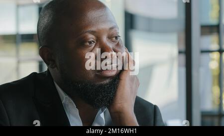 Sad unhappy dreamy afro american overweight plus size bearded man businessman feel boredom lack of motivation sit in office look at distance dream abo Stock Photo