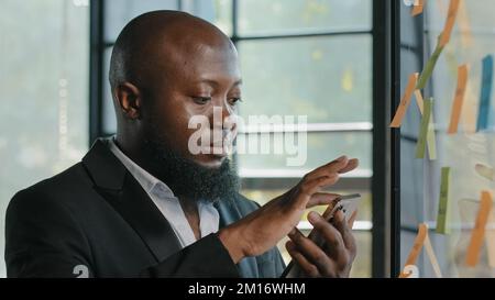 African american businessman stand near sticker board read news on smartphone make online order booking delivery at office use business mobile apps sc Stock Photo