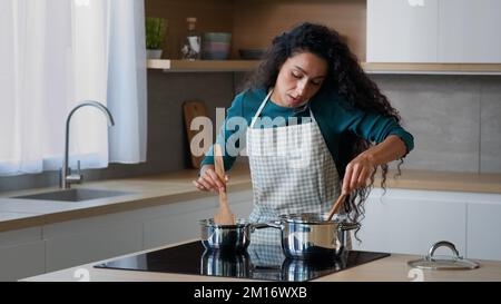 Busy arabian mom attractive curly housewife chef wears apron cooking breakfast at home kitchen stirring meal in saucepans with spoon preparing food fo Stock Photo