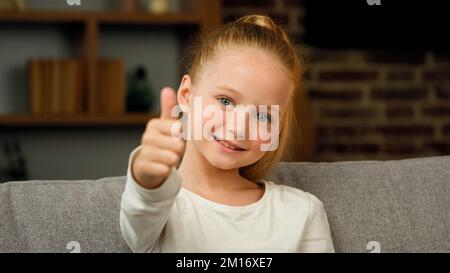 Portrait caucasian happy satisfied blonde girl little positive child kid looking at camera make thumb up like and approval gesture everything fine pos Stock Photo