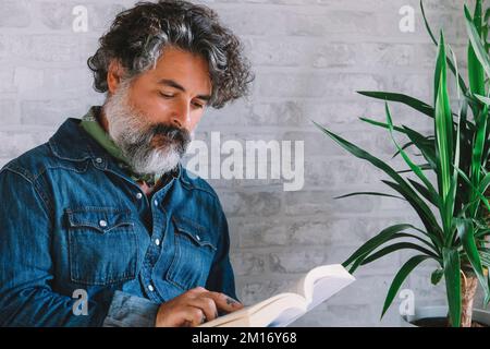A handsome, bearded man wearing a denim shirt sitting on a chair, reading a book and relaxing Stock Photo