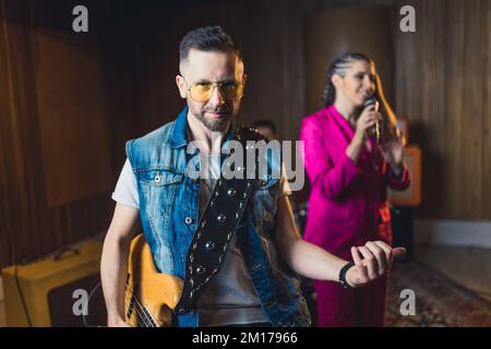 medium shot of a young bass guitarist man with yellow sunglasses, a young woman in a pink suit singing in the background. High quality photo Stock Photo