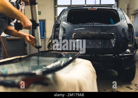 Professional unrecognizable caucasian mechanic in gray uniform using glue gun onto removed rear window glass from a black car. Blurred foreground and focus on the background. High quality photo Stock Photo