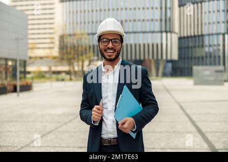 Indian construction architect in suit and hardhat with paper folder in hands against modern building Stock Photo