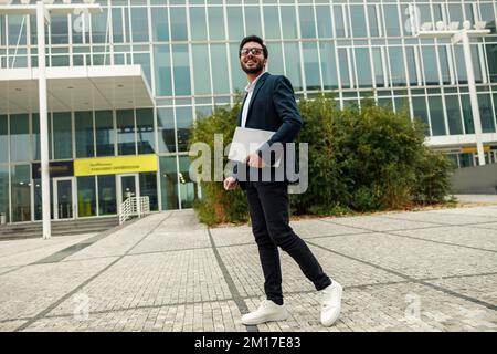 Smiling indian businessman in suit and glasses with laptop near office building Stock Photo