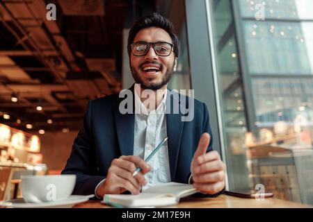 Indian businessman is making notes in notepad during working day in cafe and looking at camera Stock Photo