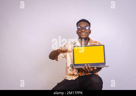 Excited young African American man sitting isolated on white wall holding laptop computer pointing to the screen Stock Photo