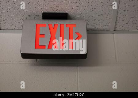 Exit sign in US school building with painted wall and drop down ceiling. EXIT lit in bright red with directional arrow pointing to the right. Stock Photo