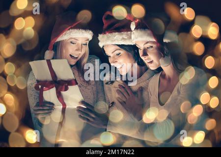 group of female students in costumes of Santa Claus opening a box of Christmas gifts. Beautiful bokeh effect from a garland. Stock Photo