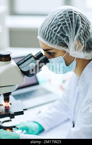 Vertical side view portrait of young female technician looking in microscope while doing research Stock Photo