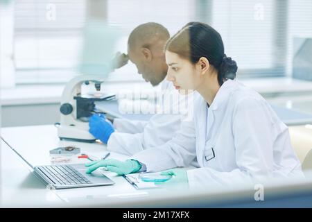 Side view portrait of Middle Eastern young woman working in medical laboratory behind glass wall and using laptop Stock Photo