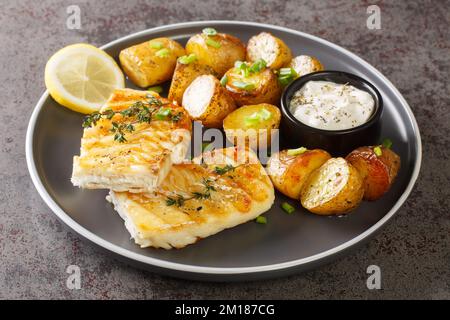 Grilled fish cod fillet with thyme served with baked potatoes, cream sauce and lemon on the table. Horizontal Stock Photo