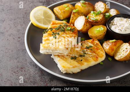 Close-up dish of grilled codfish with baked jacket potatoes, creamy sauce and lemon on the table. Horizontal Stock Photo