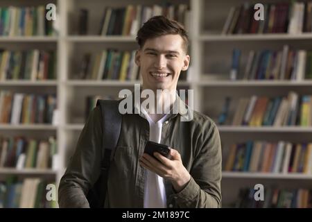Happy handsome college student guy holding smartphone, posing in library Stock Photo