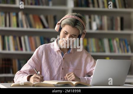 Focused student guy in pink wireless headphones studying in library Stock Photo