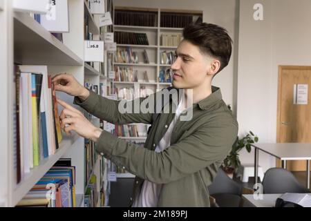 Positive handsome student guy taking textbook from bookshelf for studying Stock Photo