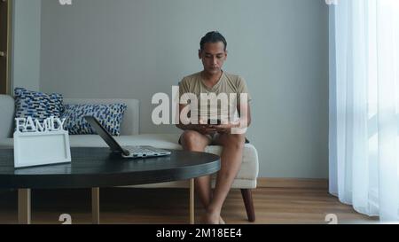 Man is using smartphone and laptop or notebook while working in the living room of the house Stock Photo