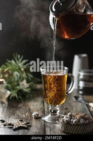 Tea is poured into a glass from a glass teapot, pouring process with steaming and Christmas decoration on wooden table. Winter time. Vertical orientat Stock Photo