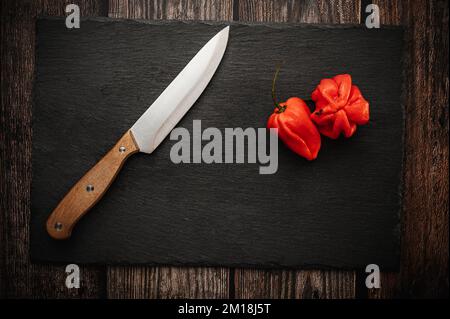 Red hot chili Habanero peppers on a slate cutting board on a dark wooden table with a knife. Top down overhead view. Stock Photo