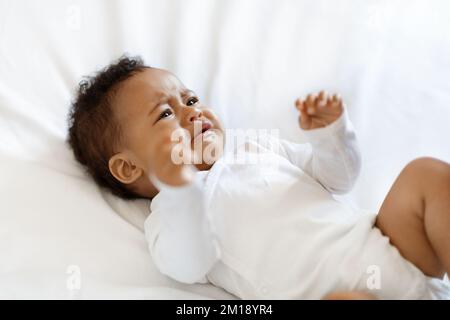 Portrait Of Crying Little Black Baby Wearing Bodysuit Lying On Bed Stock Photo