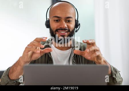 Happy latin man in headphones having video call via laptop, talking and gesturing at webcamera in home office, closeup Stock Photo
