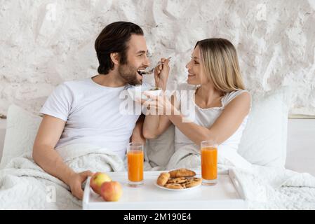 Happy loving spouses having breakfast in bed, woman feeding her husband with tasty cereal, enjoying morning together Stock Photo