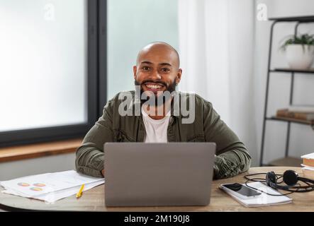 Portrait of happy latin male freelancer sitting at table, working on laptop at home office interior, copy space Stock Photo