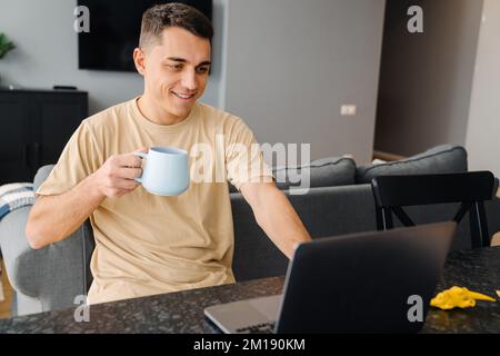 Young white man drinking tea and using laptop while sitting over table at home Stock Photo