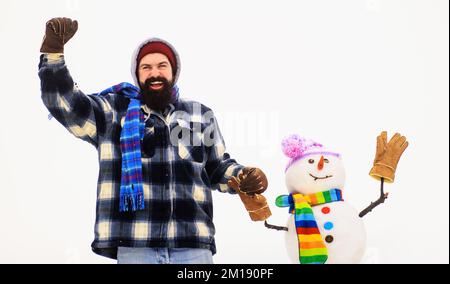 Bearded man in winter clothes holds snowman by hand. Winter fashion. Snow-man in hat, scarf, gloves. Stock Photo