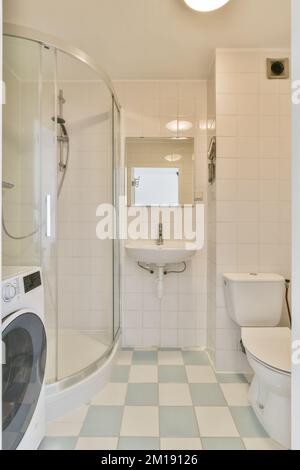 a white and blue tiled bathroom with a washer, toilet, and washing machine in the photo is taken Stock Photo