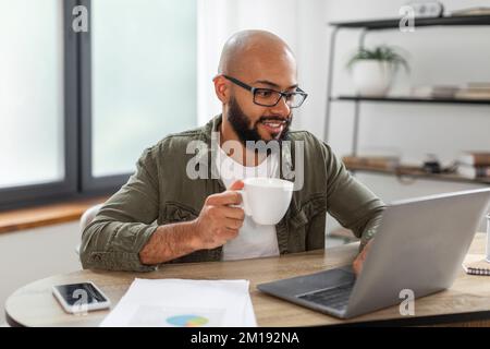 Positive latin bearded man using laptop, holding cup and enjoying drinking morning coffee while working from home Stock Photo