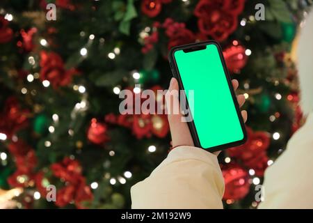 over the shoulder of hand holding green screen phone beside Christmas tree at night Stock Photo