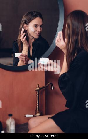 Young woman in front of the mirror putting cream on her face. Stock Photo