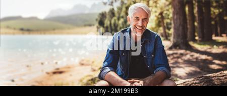 Portrait of a happy mature man sitting near a lake looking at camera and smiling. The senior caucasian man is sitting on a log by the lake on a summer Stock Photo