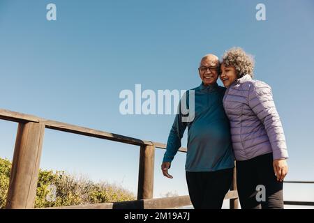 Mature couple smiling and having a good time while standing on a seaside foot bridge. Retired elderly couple spending some quality time together durin Stock Photo