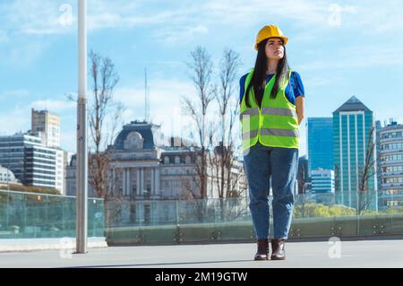 young caucasian woman engineer with safety helmet and vest, standing on the street supervising the construction site, looking to the side, with the ci Stock Photo