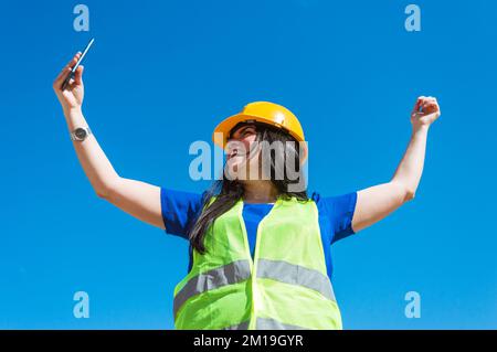 Happy young engineer woman is celebrating her new contract, raising her hands in the air smiling and looking at the phone she is holding with her hand Stock Photo