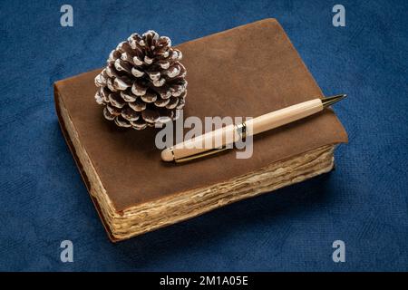 retro leather-bound journal with decked edge handmade paper pages, decorative frosty pine cone and a stylish pen, journaling and winter holidays conce Stock Photo